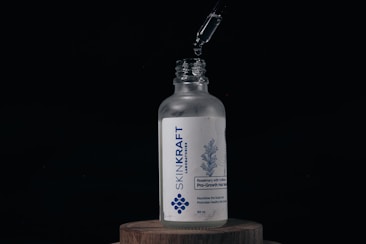 A glass bottle labeled 'SKINKRAFT LABORATORIES' stands on a wooden surface. Above the bottle, a dropper releases a droplet of liquid into the open neck. The label features a blue design of rosemary and mentions 'Rosemary with Caffeine Pro-Growth Hair Serum' in white and blue colors.