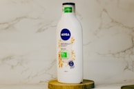 A white bottle of Nivea Naturally Good body lotion with a blue cap is placed on a wooden surface. The label indicates the product contains natural oats and is 98% natural origin ingredients. The background features a light marble surface.