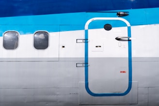 An aircraft's exterior shows part of its fuselage, including a main door and several windows. The door features a blue outline with a metal handle, and the fuselage is colored in shades of blue, white, and gray. The windows are shaped as rounded rectangles.