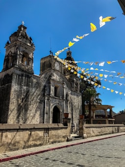A historic stone church with two bell towers and a large clock is adorned with strings of yellow and white triangular flags. The sky is clear and blue, enhancing the bright atmosphere. The church is surrounded by a cobblestone street and a low stone wall, with a palm tree visible nearby.