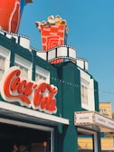 A building with a vintage cinema theme, featuring a large Coca-Cola logo sign in red script. On the roof, there is an oversized cup of Coca-Cola filled with popcorn. The facade includes decor resembling film reels, adding to the cinematic atmosphere. A marquee on the side lists items like nachos, palomitas, and hot dogs.