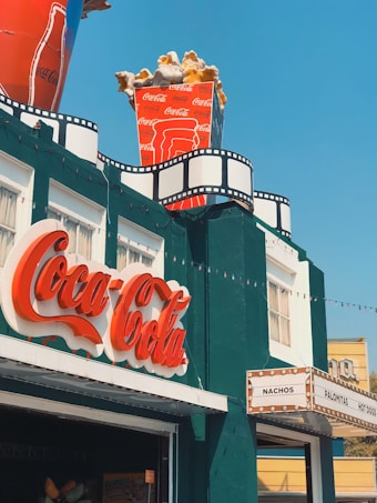 A building with a vintage cinema theme, featuring a large Coca-Cola logo sign in red script. On the roof, there is an oversized cup of Coca-Cola filled with popcorn. The facade includes decor resembling film reels, adding to the cinematic atmosphere. A marquee on the side lists items like nachos, palomitas, and hot dogs.