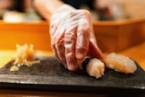 A gloved hand is delicately placing sushi, featuring a shrimp topping, onto a dark serving platter. The background is softly blurred, focusing attention on the sushi, with pickled ginger placed beside it.