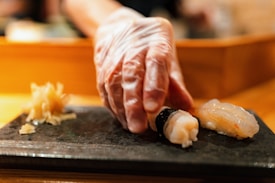A gloved hand is delicately placing sushi, featuring a shrimp topping, onto a dark serving platter. The background is softly blurred, focusing attention on the sushi, with pickled ginger placed beside it.