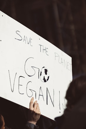A close-up of a protest sign held by a person, featuring handwritten text that reads 'SAVE THE PLANET GO VEGAN' with a small globe illustration within the text. The focus is on the sign, with the person's hand visible holding it.