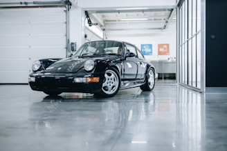 A classic black sports car is parked indoors on a shiny, polished concrete floor. The car is well-maintained and reflects the gloss from the floor and surrounding surfaces. The garage has a modern, clean look with a white roll-up door and large glass panels on the right. A metal sink or counter with blue and orange posters is visible in the background.