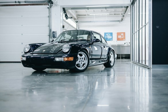 A classic black sports car is parked indoors on a shiny, polished concrete floor. The car is well-maintained and reflects the gloss from the floor and surrounding surfaces. The garage has a modern, clean look with a white roll-up door and large glass panels on the right. A metal sink or counter with blue and orange posters is visible in the background.