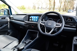 The interior of a car with a focus on the driver's area, featuring a modern steering wheel with a logo in the center, a sleek dashboard with a digital display showing several app icons, and minimalistic controls.