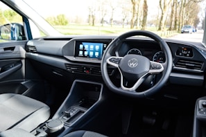 The interior of a car with a focus on the driver's area, featuring a modern steering wheel with a logo in the center, a sleek dashboard with a digital display showing several app icons, and minimalistic controls.
