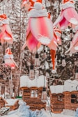 A festive outdoor scene featuring large, pink flower-shaped lanterns adorned with snow-covered Santa hats, hanging among snow-dusted trees and rustic wooden cabins. Vintage-style light bulbs are suspended between the decorations.