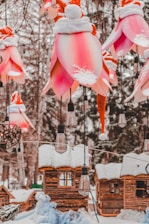 A festive outdoor scene featuring large, pink flower-shaped lanterns adorned with snow-covered Santa hats, hanging among snow-dusted trees and rustic wooden cabins. Vintage-style light bulbs are suspended between the decorations.