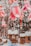 A festive outdoor scene featuring large, pink flower-shaped lanterns adorned with snow-covered Santa hats, hanging among snow-dusted trees and rustic wooden cabins. Vintage-style light bulbs are suspended between the decorations.