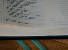 A close-up view of an open book on a wooden table, showing text and some footnotes. A teal ribbon bookmark is visible protruding from the pages.