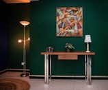 A modern interior space with a deep green wall, featuring a colorful abstract painting with geometric patterns and hands. Below the painting, a sleek wooden console table with metallic legs holds a potted plant and a decorative lamp with a newspaper print shade. To the left, a tall floor lamp with three lights illuminates the corner, which includes a round woven rug on a polished floor.