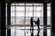 Silhouetted figures of two construction workers, wearing hard hats, stand inside a building framework, engaged in observing blueprints. The large window in the background offers a view of an overcast sky and distant houses. The floor reflects the scaffolding and figures, suggesting recent rainfall or a water spill.
