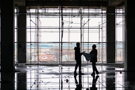 Silhouetted figures of two construction workers, wearing hard hats, stand inside a building framework, engaged in observing blueprints. The large window in the background offers a view of an overcast sky and distant houses. The floor reflects the scaffolding and figures, suggesting recent rainfall or a water spill.