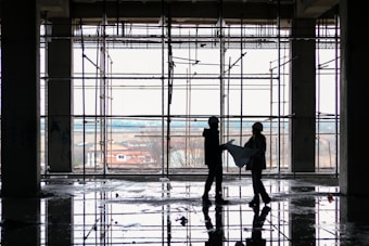 Silhouetted figures of two construction workers, wearing hard hats, stand inside a building framework, engaged in observing blueprints. The large window in the background offers a view of an overcast sky and distant houses. The floor reflects the scaffolding and figures, suggesting recent rainfall or a water spill.