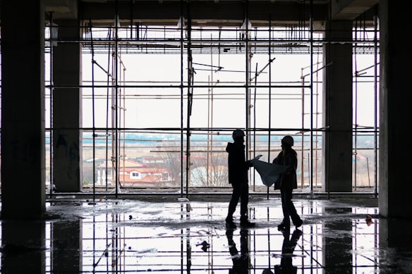 Silhouetted figures of two construction workers, wearing hard hats, stand inside a building framework, engaged in observing blueprints. The large window in the background offers a view of an overcast sky and distant houses. The floor reflects the scaffolding and figures, suggesting recent rainfall or a water spill.