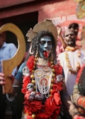 A person is dressed in elaborate traditional attire resembling the Hindu goddess Kali. The face is painted blue with white designs, and the person is adorned with heavy jewelry including garlands with skulls and beads. The tongue is sticking out in a fierce expression, and they hold a large curved prop in their hand. The background includes other people, some of whom are also adorned with garlands.