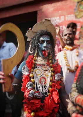 A person is dressed in elaborate traditional attire resembling the Hindu goddess Kali. The face is painted blue with white designs, and the person is adorned with heavy jewelry including garlands with skulls and beads. The tongue is sticking out in a fierce expression, and they hold a large curved prop in their hand. The background includes other people, some of whom are also adorned with garlands.