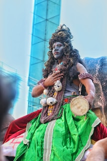 A person is dressed in vibrant and elaborate traditional attire, featuring a bright green skirt, decorative beads, and a painted face. They are holding a small drum and seem to be part of a cultural or religious festivity. The background shows a modern building structure, juxtaposing traditional and contemporary elements.