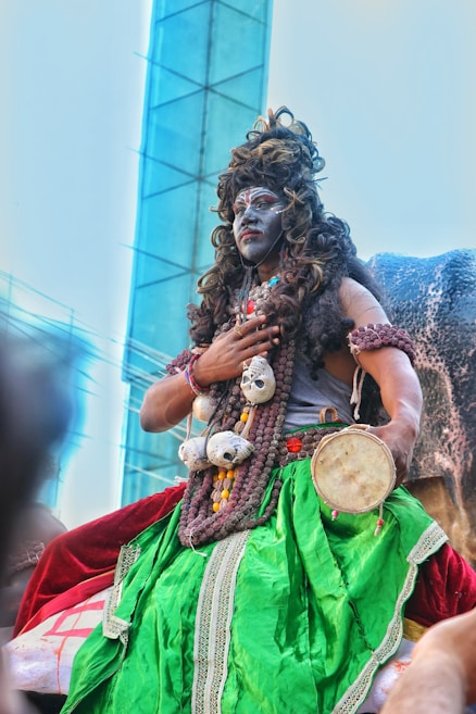 A person is dressed in vibrant and elaborate traditional attire, featuring a bright green skirt, decorative beads, and a painted face. They are holding a small drum and seem to be part of a cultural or religious festivity. The background shows a modern building structure, juxtaposing traditional and contemporary elements.