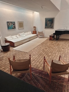 A modern living room with a minimalist design features a long white sofa against an off-white wall. The room includes artworks hanging on the walls and two fabric armchairs facing a black grand piano. The floor is covered with light brown tiles, and there is a Persian-style rug near the chairs, adding a touch of warmth. Natural light softly illuminates the space, highlighting a sculpture in the corner and emphasizing the clean, open layout.