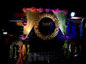 A decorative backdrop features a circle made of flowers with a word written in a script, surrounded by golden drapes and pink flowers at the top. There are floral arrangements on either side, and two decorative umbrellas flanking the setup. The lighting is dramatic, highlighting the floral and fabric elements.