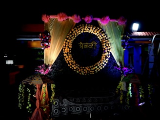 A decorative backdrop features a circle made of flowers with a word written in a script, surrounded by golden drapes and pink flowers at the top. There are floral arrangements on either side, and two decorative umbrellas flanking the setup. The lighting is dramatic, highlighting the floral and fabric elements.
