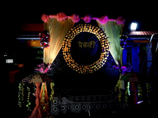 A decorative backdrop features a circle made of flowers with a word written in a script, surrounded by golden drapes and pink flowers at the top. There are floral arrangements on either side, and two decorative umbrellas flanking the setup. The lighting is dramatic, highlighting the floral and fabric elements.