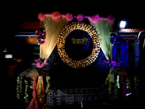 A decorative backdrop features a circle made of flowers with a word written in a script, surrounded by golden drapes and pink flowers at the top. There are floral arrangements on either side, and two decorative umbrellas flanking the setup. The lighting is dramatic, highlighting the floral and fabric elements.