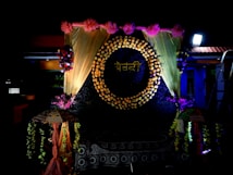 A decorative backdrop features a circle made of flowers with a word written in a script, surrounded by golden drapes and pink flowers at the top. There are floral arrangements on either side, and two decorative umbrellas flanking the setup. The lighting is dramatic, highlighting the floral and fabric elements.