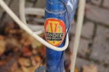 A close-up view of a bicycle frame featuring a vintage VINDEC badge in red and yellow with black text. The blue frame has white cables running across it and is surrounded by a background of blurred autumn leaves and cobblestones.