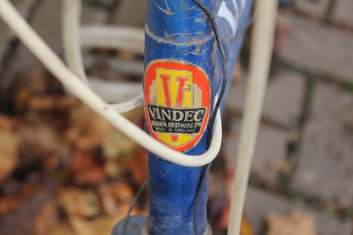 A close-up view of a bicycle frame featuring a vintage VINDEC badge in red and yellow with black text. The blue frame has white cables running across it and is surrounded by a background of blurred autumn leaves and cobblestones.