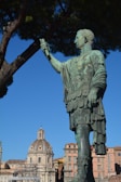 A large bronze statue of a Roman figure is prominently featured, standing in an authoritative pose with an arm raised. The background includes historical architecture, such as a domed building and several multi-story structures. A tall tree partially frames the image, with branches extending overhead.