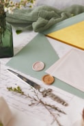 A collection of stationery items including envelopes, wax seals, and a letter opener are arranged on a surface. The scene is accented with greenery and delicate flowers in a green glass vase. The envelopes are pastel shades of mint and pink, and there is a bright yellow card peeking out. The wax seals are ornate, featuring floral designs.