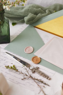 A collection of stationery items including envelopes, wax seals, and a letter opener are arranged on a surface. The scene is accented with greenery and delicate flowers in a green glass vase. The envelopes are pastel shades of mint and pink, and there is a bright yellow card peeking out. The wax seals are ornate, featuring floral designs.
