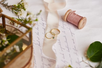 Two gold rings with intricate designs sit atop a handwritten letter. Nearby, there is a spool of pink ribbon, lush greenery, and small white flowers. A brass and glass box with floral accents is partially visible.