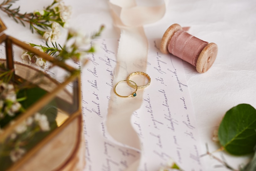 Two gold rings with intricate designs sit atop a handwritten letter. Nearby, there is a spool of pink ribbon, lush greenery, and small white flowers. A brass and glass box with floral accents is partially visible.