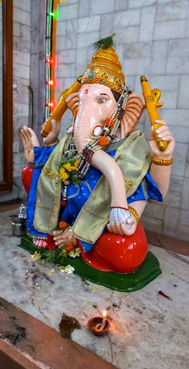 A colorful statue of the Hindu deity Ganesha is adorned with traditional decorations and flowers. The statue is placed on a marble surface with a small lit oil lamp in front. Multicolored lights are visible in the background, creating a festive atmosphere.