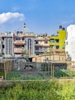 A series of colorful residential buildings with ornate architectural details, including arched windows and decorative patterns, are prominently displayed. Several buildings are adorned with prayer flags. In the foreground, a garden or small farm with green plants and yellow flowers is enclosed by simple structures made of wood and corrugated metal.