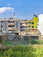 A series of colorful residential buildings with ornate architectural details, including arched windows and decorative patterns, are prominently displayed. Several buildings are adorned with prayer flags. In the foreground, a garden or small farm with green plants and yellow flowers is enclosed by simple structures made of wood and corrugated metal.