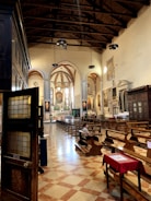 A well-lit church interior featuring wooden pews, stained glass windows, and painted artwork on the walls. The ceiling is high with exposed beams and pendant lights. A few people are seated, with one visible inside the church. The floor is tiled in a checkerboard pattern.