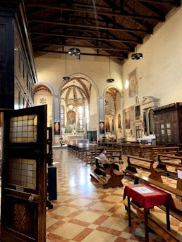 A well-lit church interior featuring wooden pews, stained glass windows, and painted artwork on the walls. The ceiling is high with exposed beams and pendant lights. A few people are seated, with one visible inside the church. The floor is tiled in a checkerboard pattern.