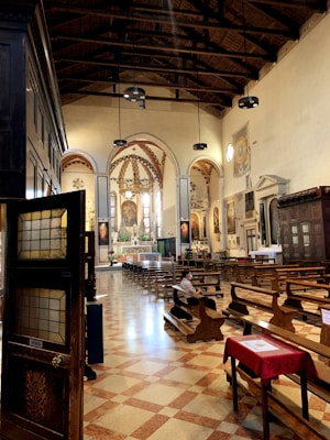 A well-lit church interior featuring wooden pews, stained glass windows, and painted artwork on the walls. The ceiling is high with exposed beams and pendant lights. A few people are seated, with one visible inside the church. The floor is tiled in a checkerboard pattern.