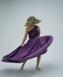A woman is spinning, causing her long purple skirt to flare out in motion. She wears a sleeveless top with sparkling details and beige high heels. Her blonde hair is styled in loose curls, adding to the dynamic movement.