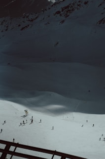 A snowy mountain slope with a group of people skiing. The scene is set against a backdrop of rugged, dark mountain terrain. The snow is bright under the sunlight, creating a strong contrast with the shadowed areas of the mountain. Ski tracks are visible in the snow, and a few ski signs and barriers can be seen.
