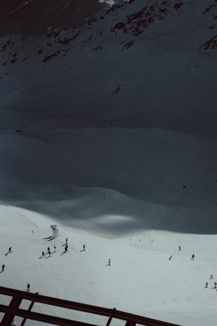A snowy mountain slope with a group of people skiing. The scene is set against a backdrop of rugged, dark mountain terrain. The snow is bright under the sunlight, creating a strong contrast with the shadowed areas of the mountain. Ski tracks are visible in the snow, and a few ski signs and barriers can be seen.