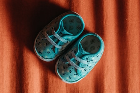 A pair of small blue baby shoes with laces lies on a surface with a brown, textured background. The shoes feature a simple pattern and appear soft and comfortable.
