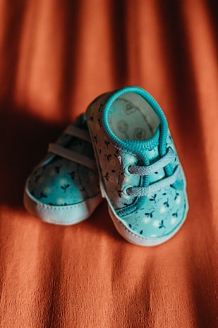 A pair of small, blue baby shoes with a floral pattern on the fabric, resting on an orange textured surface.